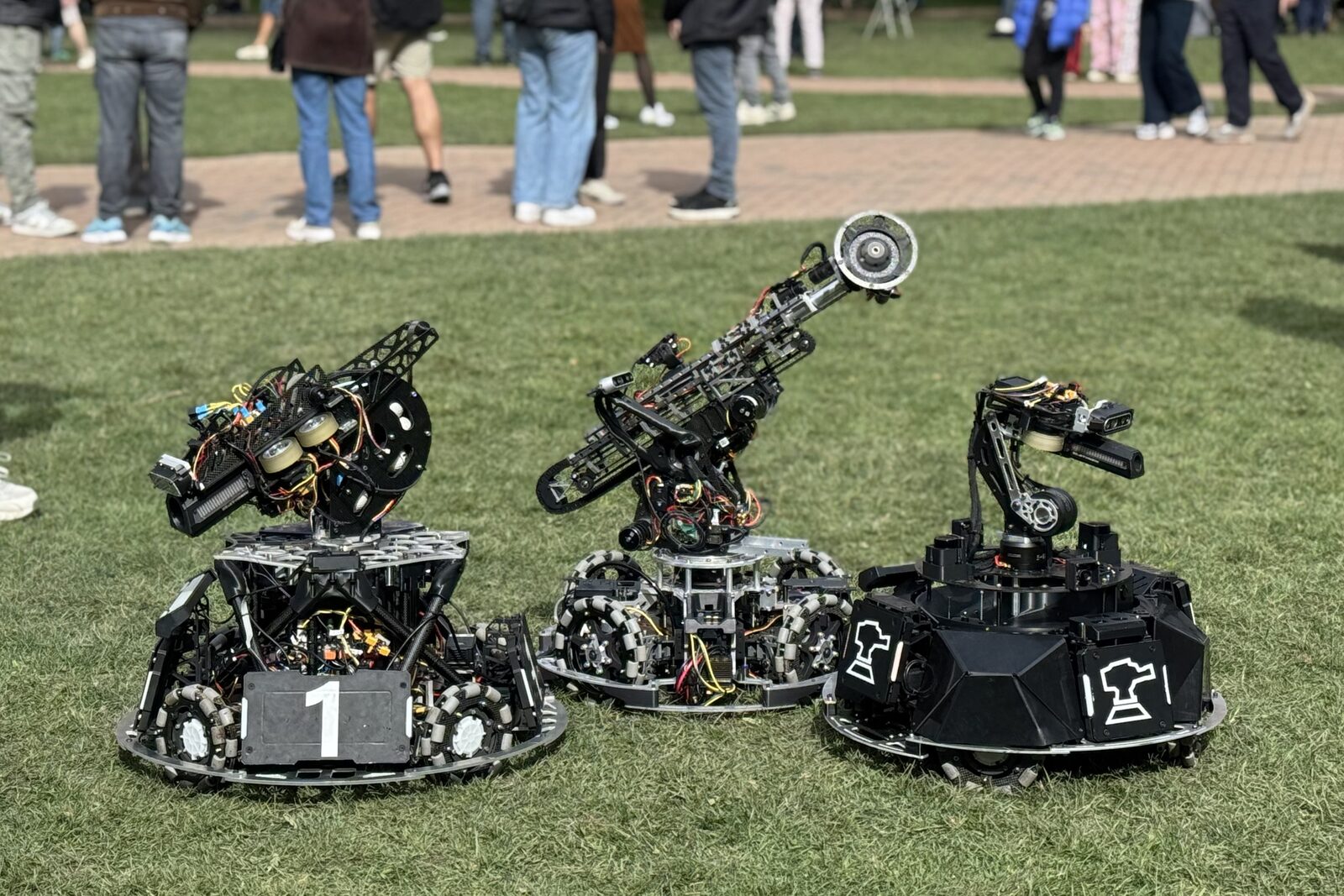 Three of ARUW's 2026 robots side by side on the grass, two with long launcher barrels and the black-shelled sentry on the right, with visitors' legs in the background