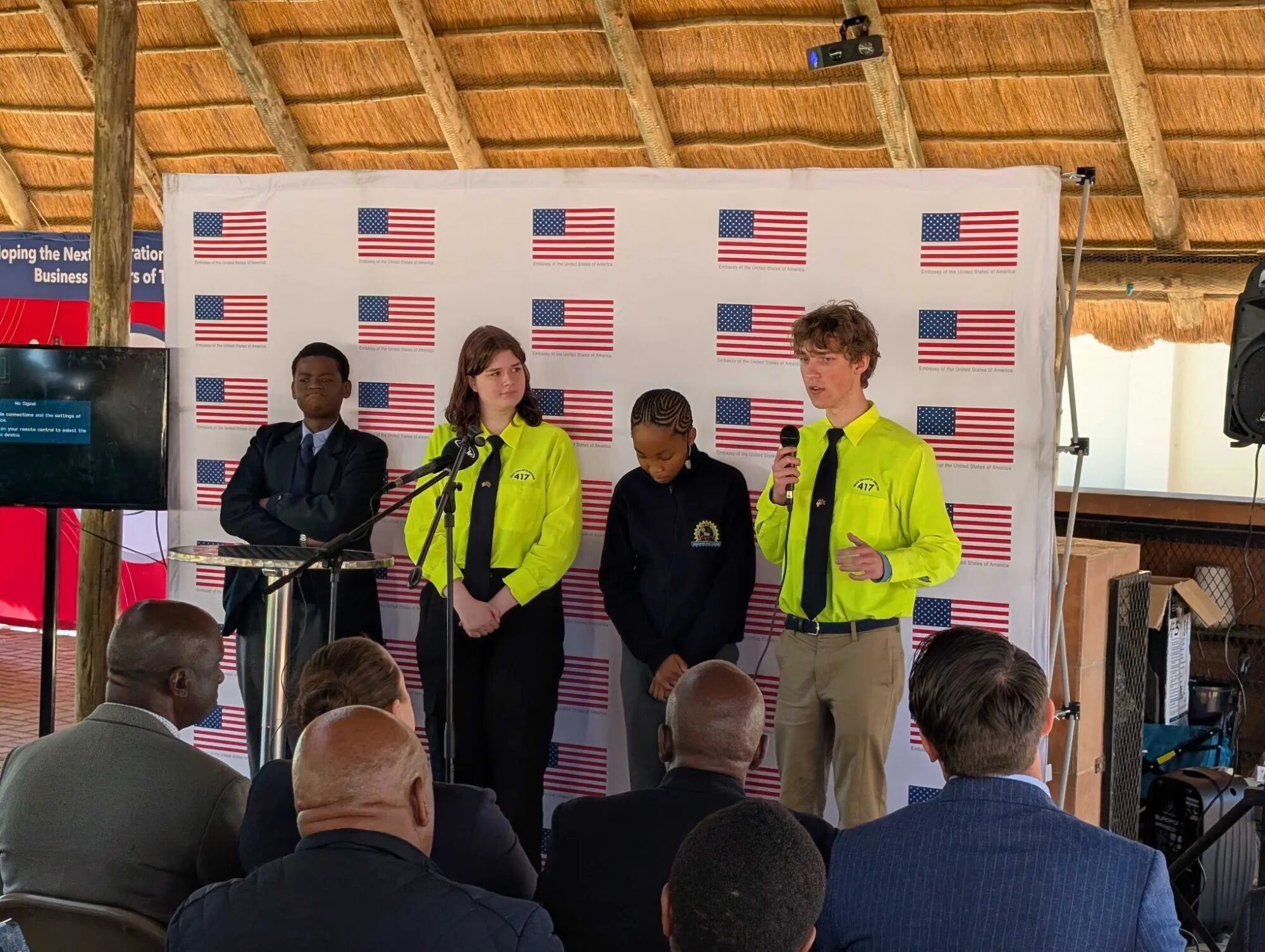 Eric speaking into a microphone in front of a backdrop of American flags, with a teammate and two Botswana students beside him