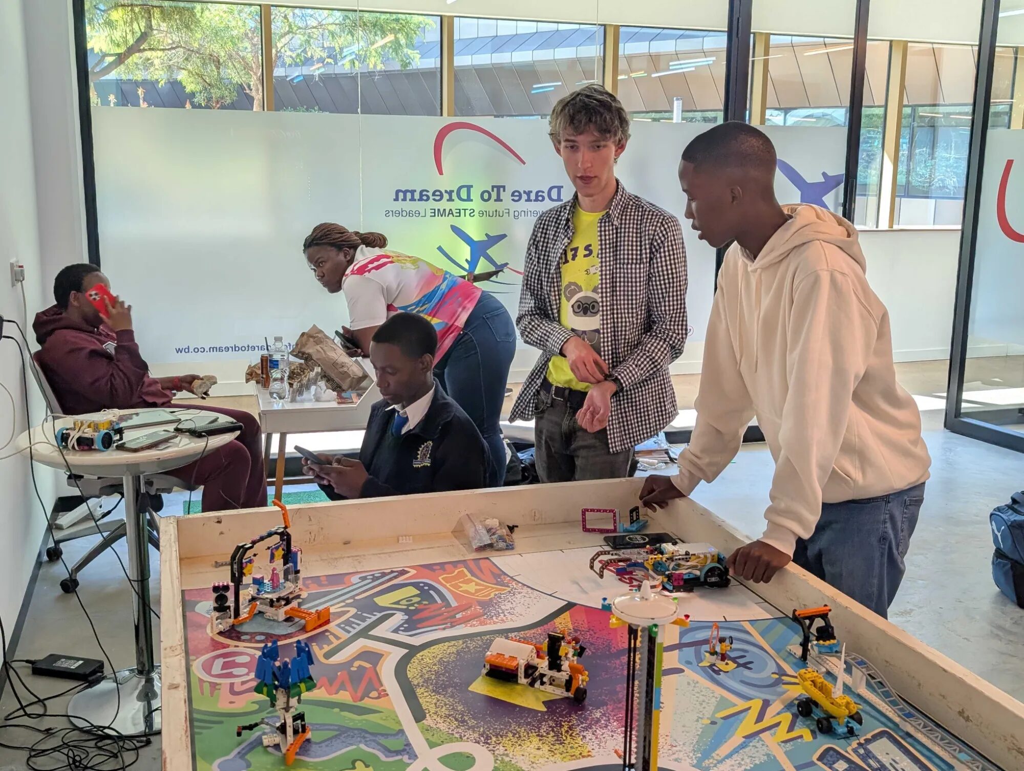 Eric and a student from the FLL team talking over a LEGO robot on a FIRST LEGO League field, with other students working behind them