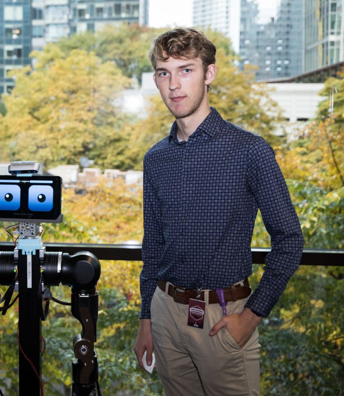Eric standing next to OpenDubs in front of a window full of autumn trees and city buildings; the robot's tablet face shows two cartoon eyes above its arms