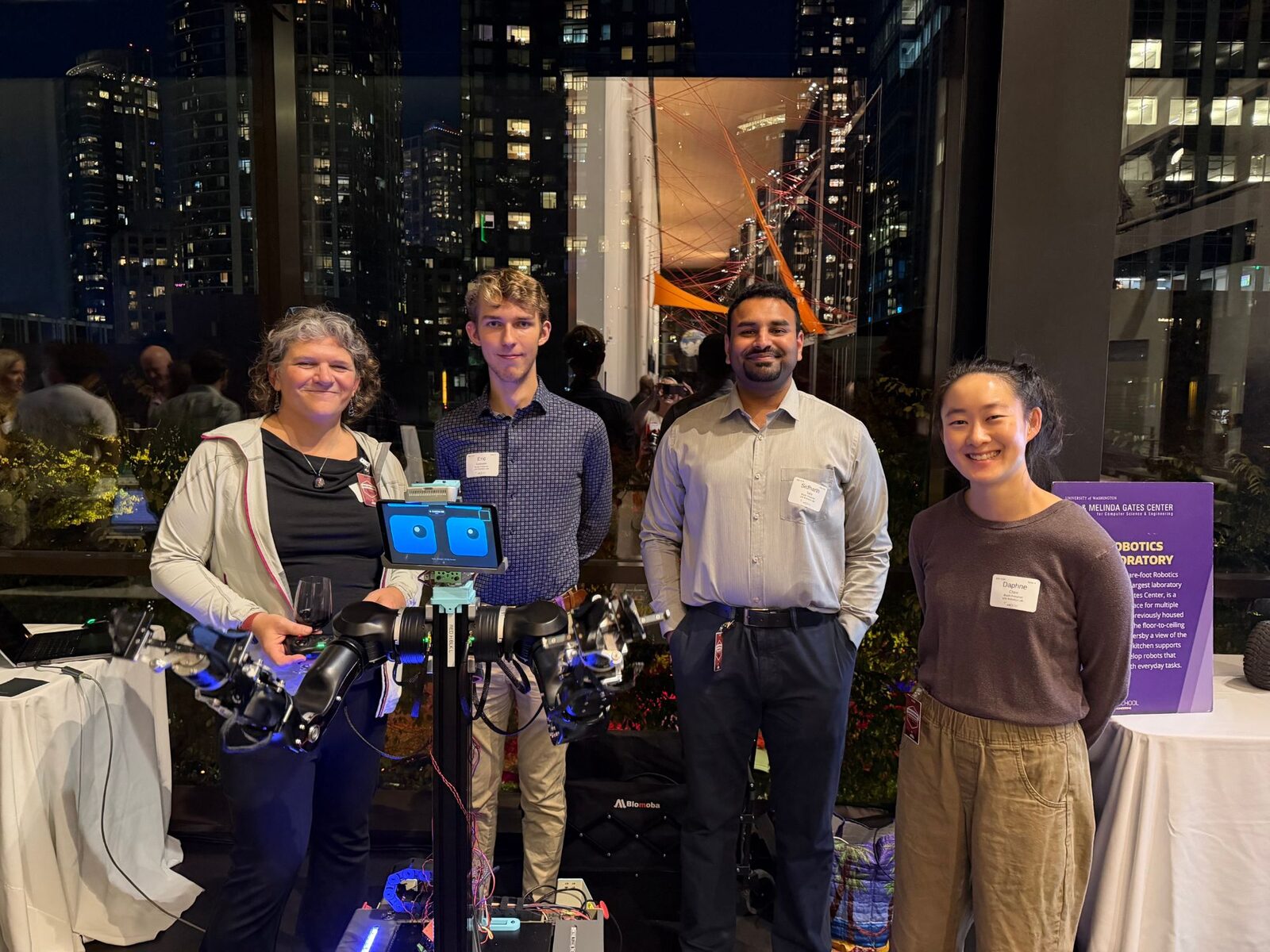 Four people standing behind OpenDubs at a night event, the robot's two arms spread out in front of them and its tablet face showing two cartoon eyes, with Seattle's lit-up towers through the windows behind them